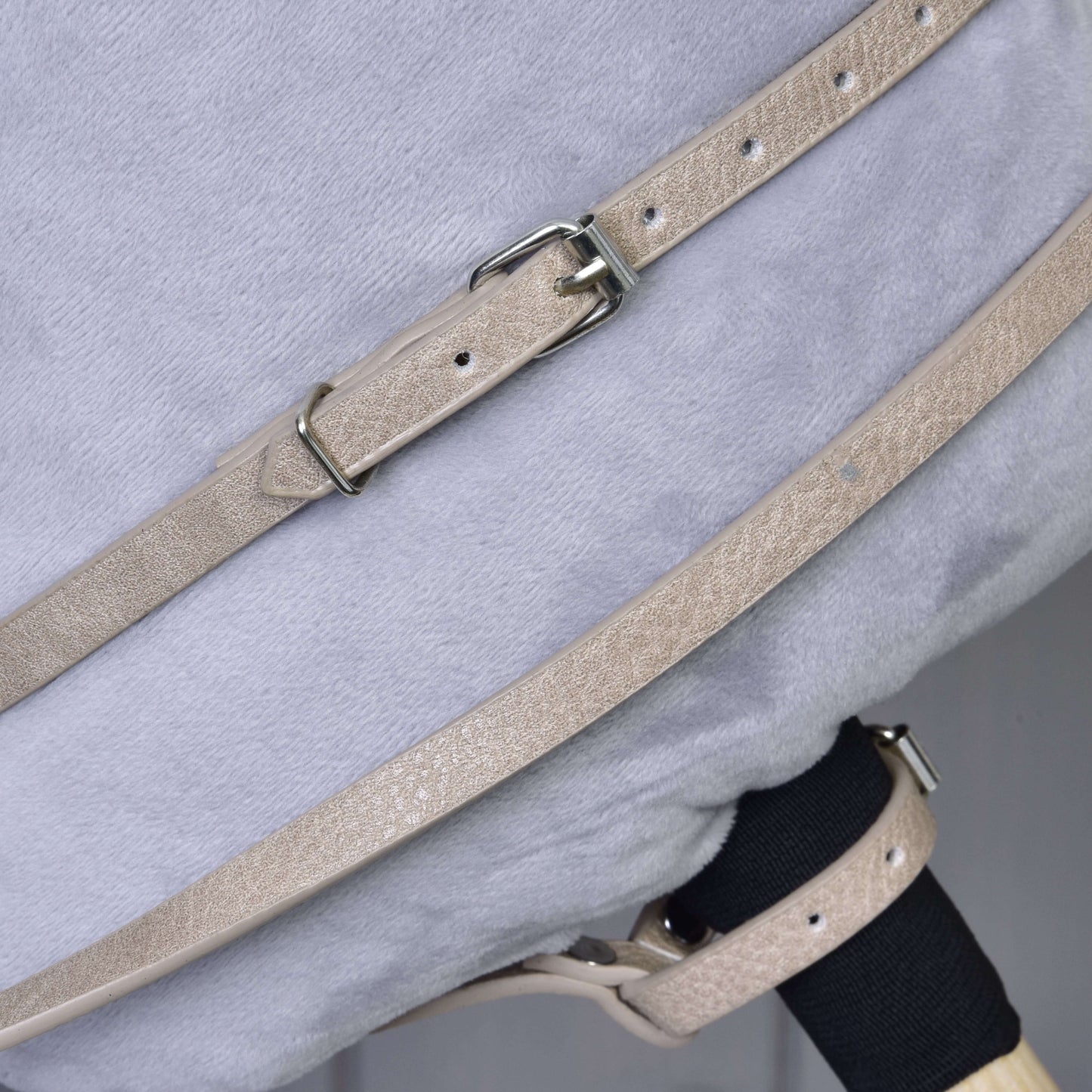 A close-up of beige Bordeaux leatherette straps with metal buckles from the Hobby Horse LarDen bridle, reins, and martingale set, fastened over a soft light gray plush surface.