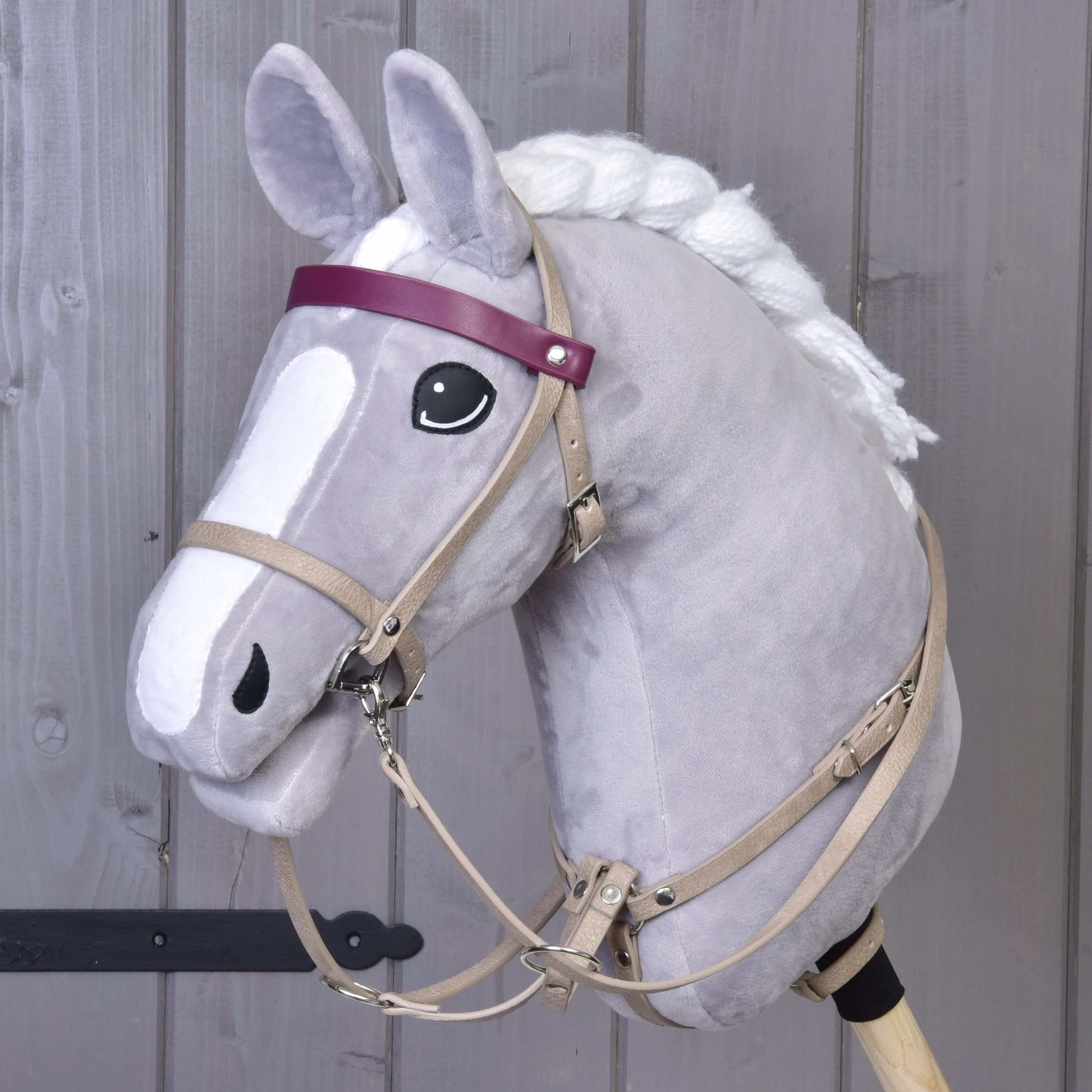 A plush gray hobby horse by Hobby Horse LarDen, featuring a Bordeaux leatherette bridle set with reins and martingale, white mane, black and white eyes, and a purple headband stands against a wooden background.