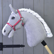 A plush gray Hobby Horse LarDen with a white braided mane, black eye, and Halter raspberry (size M), attached to a wooden stick, is set before a wooden gray paneled wall.