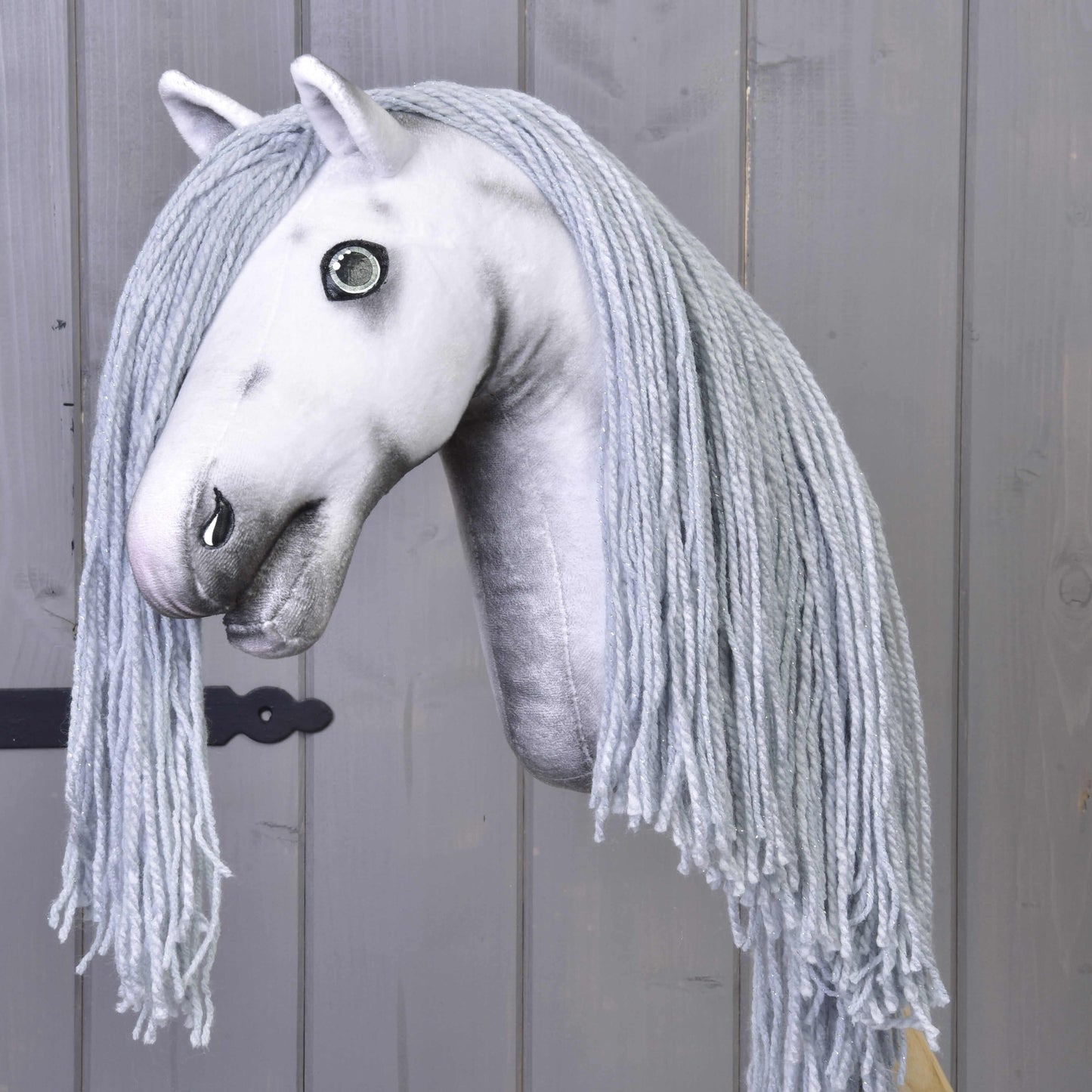 Hobby Horse Einhorn Silver mit Trense und Halfter von Hobby Horse LarDen mit weißem Plüschkopf, langer geflochtener hellgrauer Garnmähne und auf einem Stock montiert, vor grauem Holz-Hintergrund.