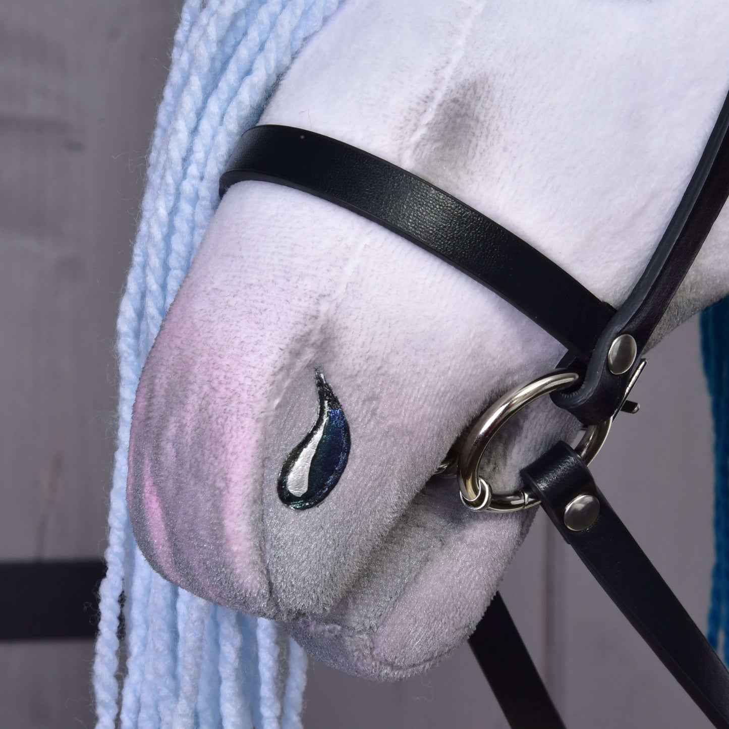 Hobby Horse Einhorn Rainbow mit Trense und Halfter von Hobby Horse LarDen, Nahaufnahme des Gesichts mit hellblauer Wollmähne, schwarzer Trense und schwarzem Halfter sowie einem tropfenförmigen Fleck nahe der Nase vor einem hellen Holzhintergrund.