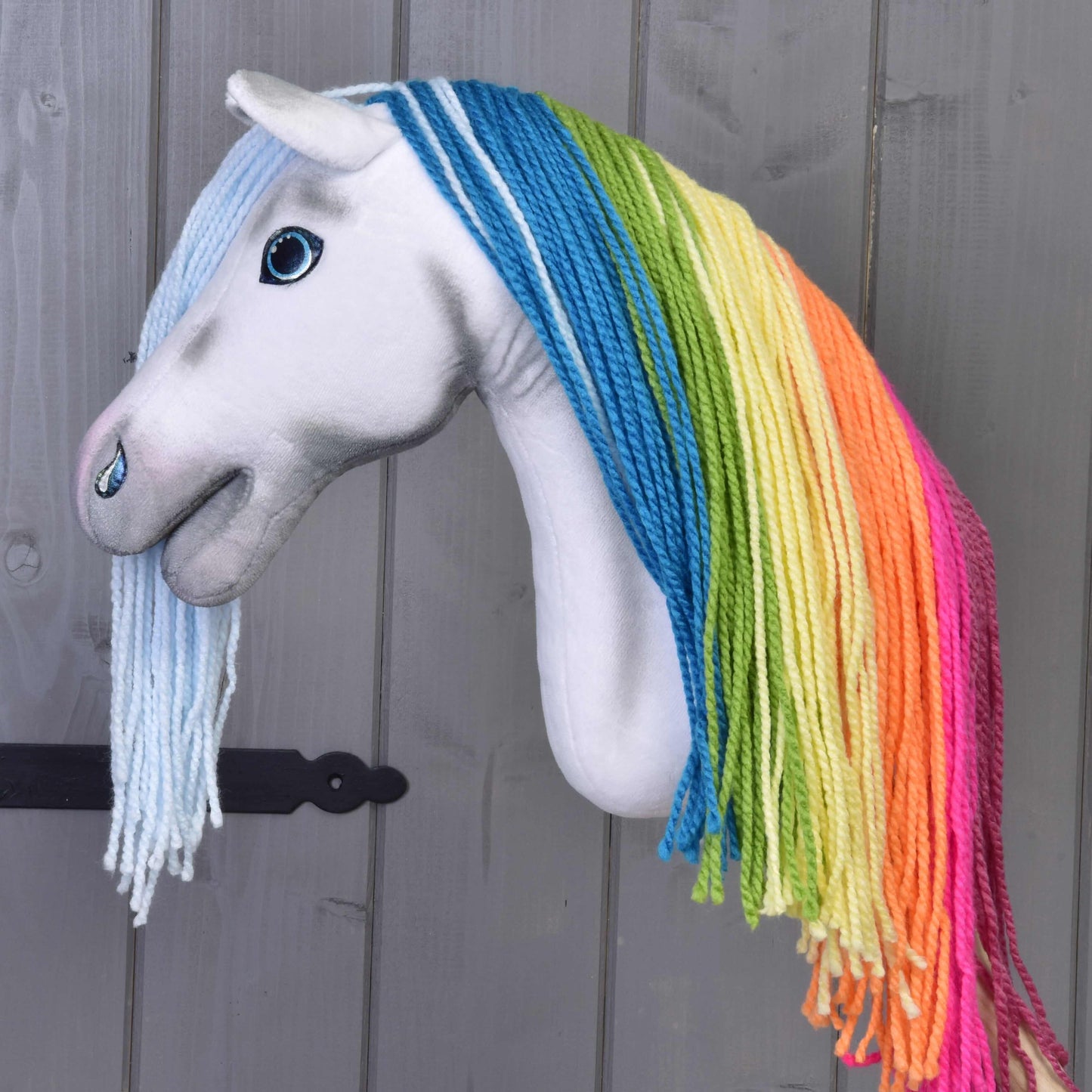 Hobby Horse LarDen „Hobby Horse Einhorn Rainbow mit Trense und Halfter“ mit weißem Gesicht und leuchtender Regenbogenmähne aus Garn vor Holzhintergrund.