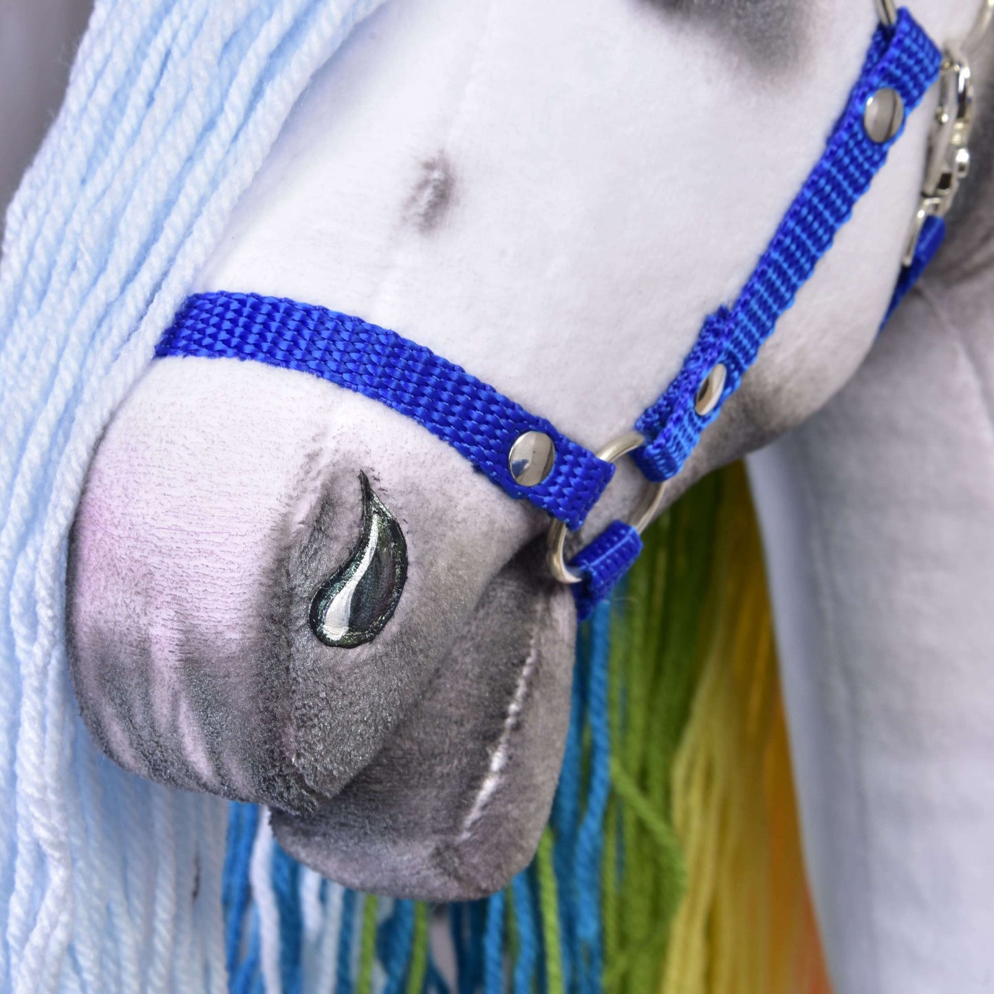 Hobby Horse Einhorn Rainbow mit Trense und Halfter von Hobby Horse LarDen in Nahaufnahme mit weißem Plüschgesicht, blauem Halfter, grau schattierter Schnauze, glänzend gestickter Nüster, hellblauer Wollmähne und grünem Schopf.