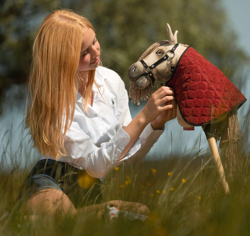 Eine junge Person mit langen blonden Haaren und weißem Shirt sitzt im hohen Gras und richtet behutsam das Zaumzeug eines Hobby Horse mit roter gesteppter Decke, vor verschwommenem Grün im Hintergrund.