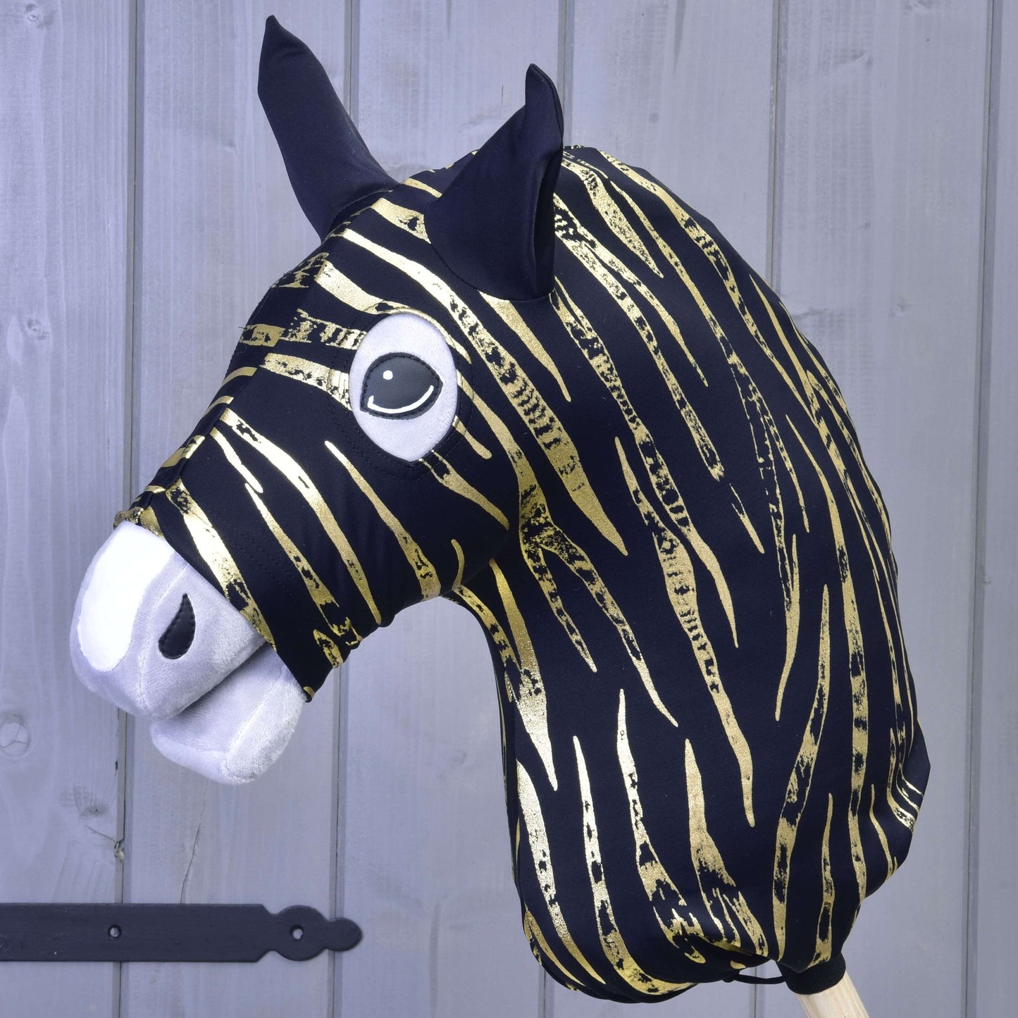 La Combinaison de protection Hobby Horse Zèbre Or (Lycra) de Hobby Horse LarDen présente une tête zébrée noire et dorée, des oreilles noires, un museau blanc en feutrine et de grands yeux cartoon, devant un mur en bois gris clair.