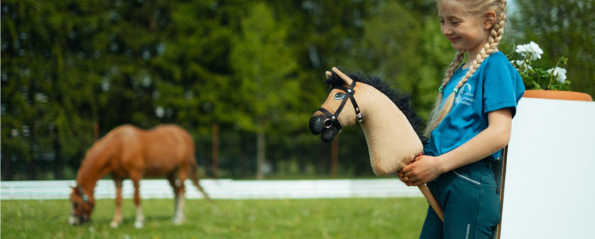 A young girl with blond braids, wearing a blue shirt and pants, smiles while hobby horsing with her toy hobby horse in a grassy field. A real horse grazes in the background near a white fence and trees.