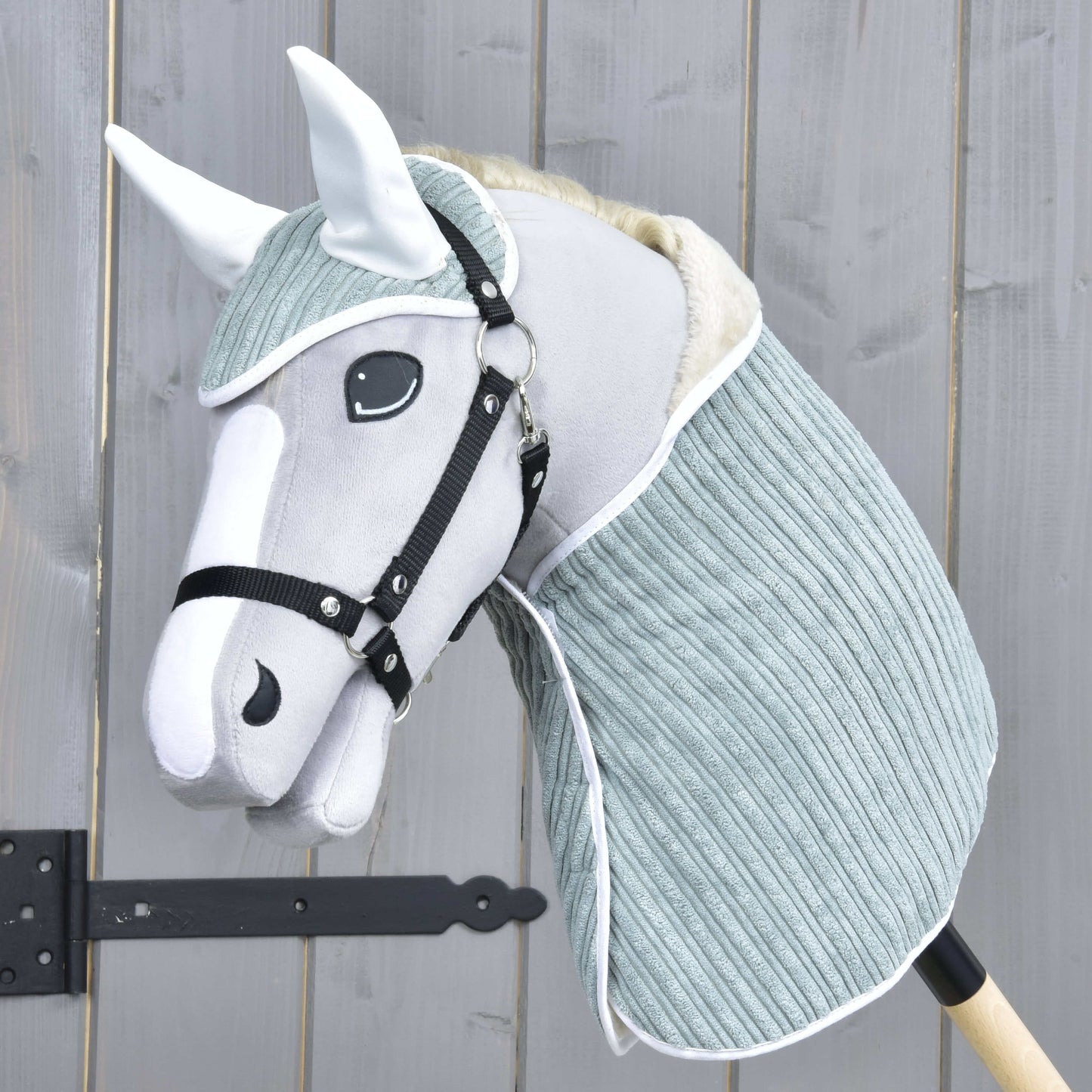 Set Manchester Little Green von Hobby Horse LarDen zeigt ein grauweißes Hobby Horse mit weichem Stoffkopf, schwarzem Zaumzeug, Fliegenhaube und hell blaugrünem Halsteil an einem Holzstab, vor einem Holzzaun.
