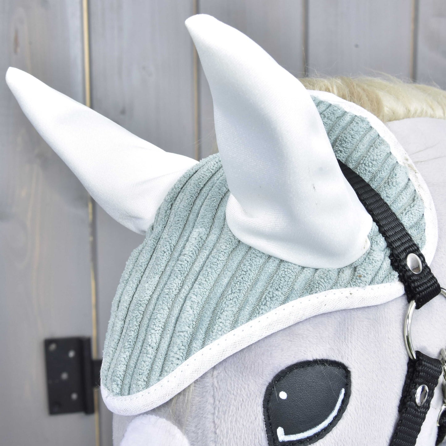 Nahaufnahme von Hobby Horse LarDen Set Manchester Little Green: ein Plüschpferdekopf mit strukturierter hellgrauer Fliegenhaube, großen weißen Ohren, schwarzem Zaumzeug und gesticktem Auge, vor einem Holzhintergrund.