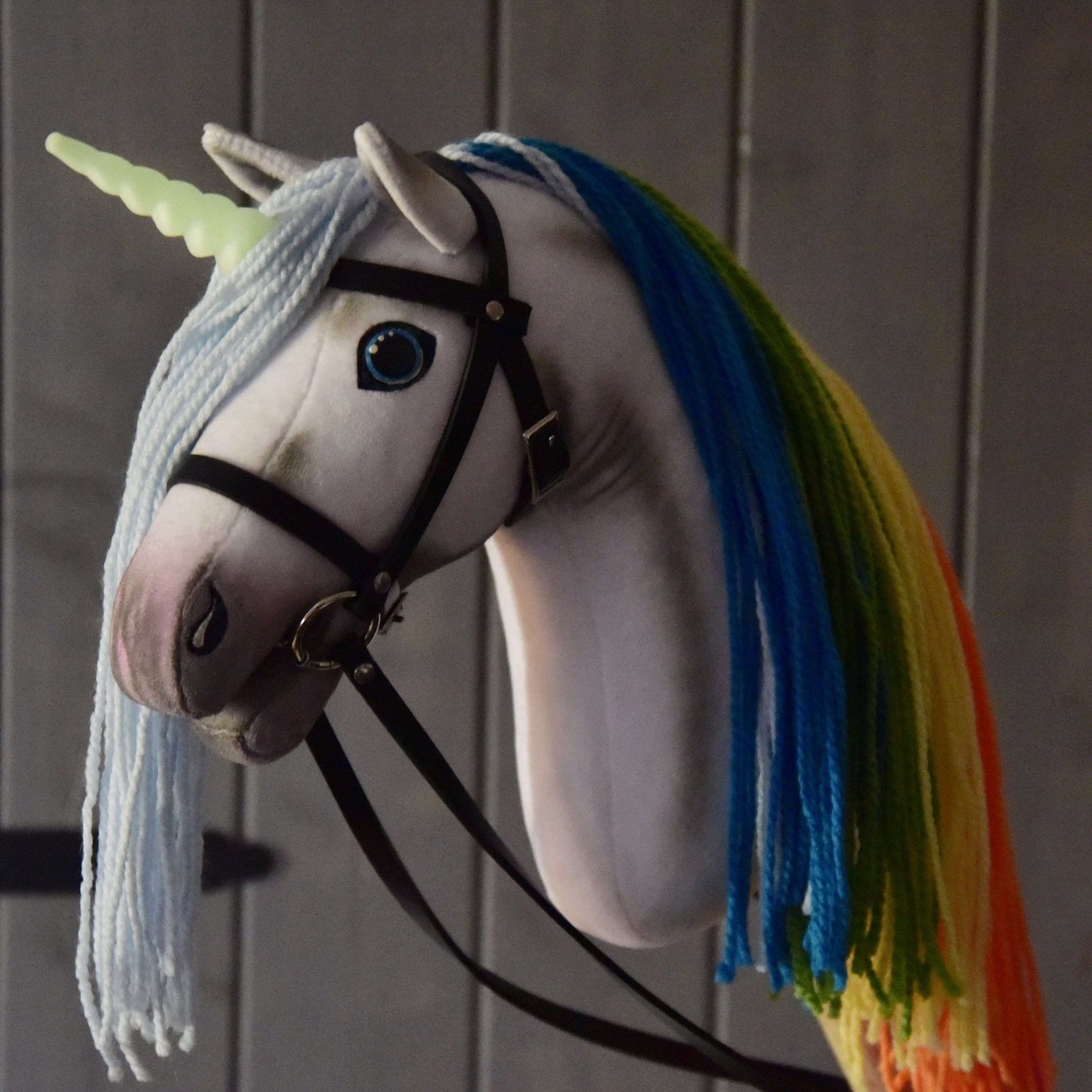 Hobby Horse LarDen Hobby Horse Einhorn Rainbow mit Trense und Halfter mit Plüsch-Einhorn, blauen Augen, blassgrünem Horn, hellblauer Trense und Regenbogenmähne aus blauem, grünem, gelbem und orangefarbenem Garn vor Holzhintergrund.