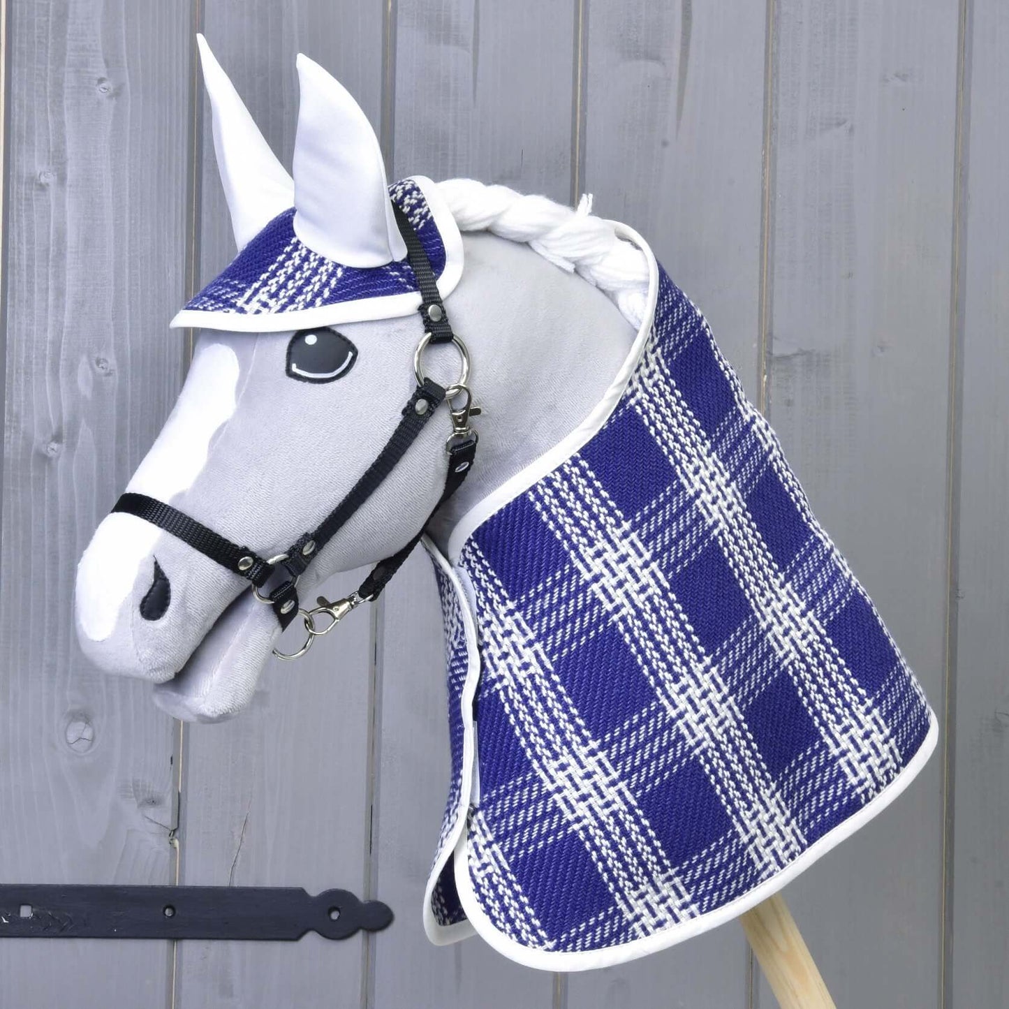Ein weißes Hobby Horse LarDen Spielzeug mit schwarzem Zaumzeug trägt die karierte Decke und Haube aus dem Set Blue Cube und steht vor einem grauen Holzhintergrund.