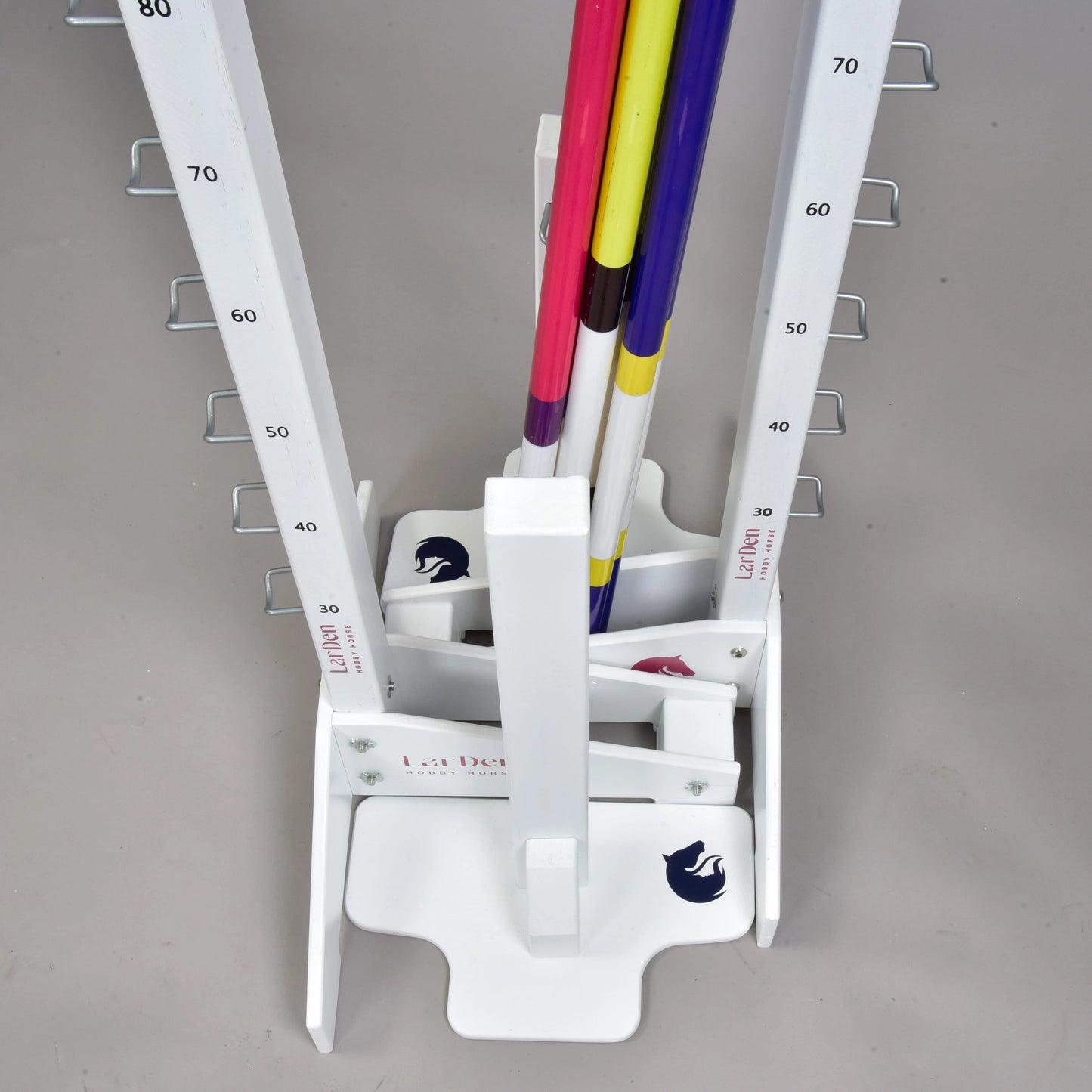 Hindernis Set für Hobby Horsing mit einem weißen senkrechten Gestell mit nummerierten Markierungen und Haken, das farbige Stangen senkrecht für verstellbare Hindernisse hält, ideal für Hobby Horsing. Die Basis zeigt ein Pferdelogo und die Marke „Hobby Horse LarDen“. Der Boden ist hellgrau.
