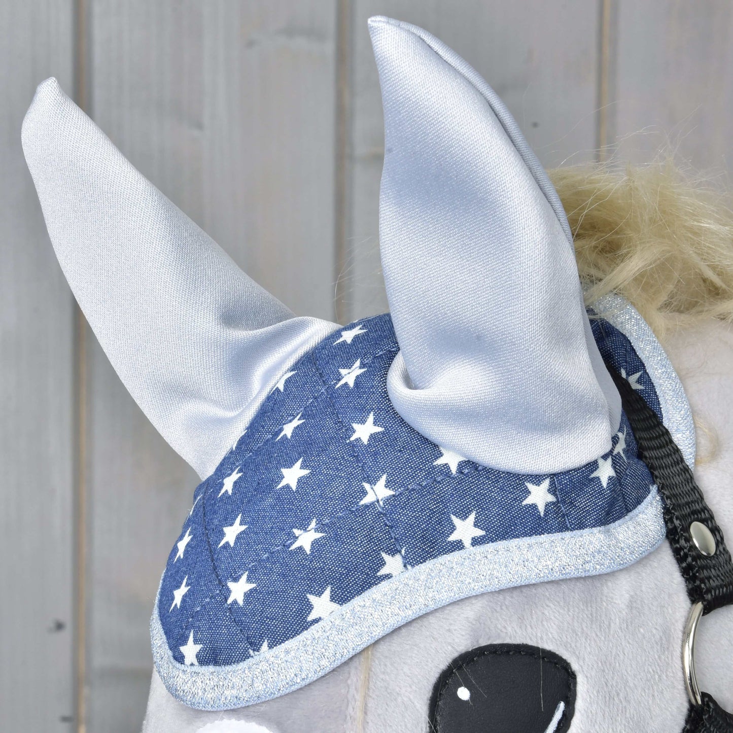 Gros plan d’un cheval portant le Bonnet d’oreilles Étoile de Hobby Horse LarDen, avec design bleu à étoiles blanches et bordure argentée pour une protection élégante contre les mouches. Une partie de l’œil du cheval est visible sur fond de panneaux en bois clair.
