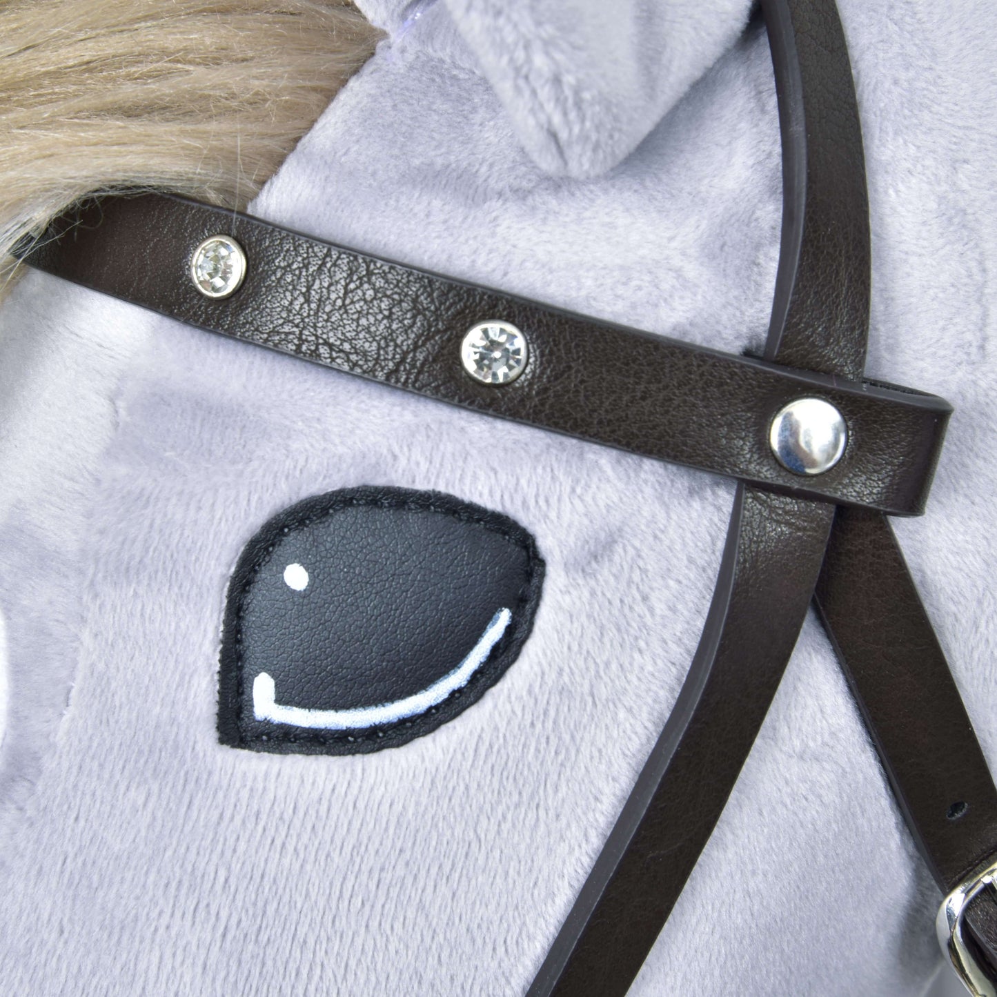 Nahaufnahme des Gesichts eines Plüsch-Spielzeugpferds mit gesticktem schwarz-weißem Auge, hellgrauem Fell und dem Hobby Horse LarDen Komplettes Kunstlederzaumzeug Braun mit Ziersteinen.
