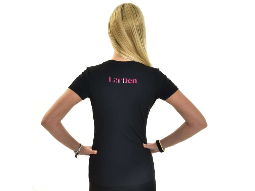 Eine Person mit langen blonden Haaren steht mit dem Rücken zur Kamera, die Hände in die Hüften gestützt, und trägt das Hobby Horse LarDen T-Shirt LarDen Active Black vor weißem Hintergrund, wobei das glatte Spandex-Material und „LarDen“ in Rosa auf dem oberen Rücken hervorgehoben werden.