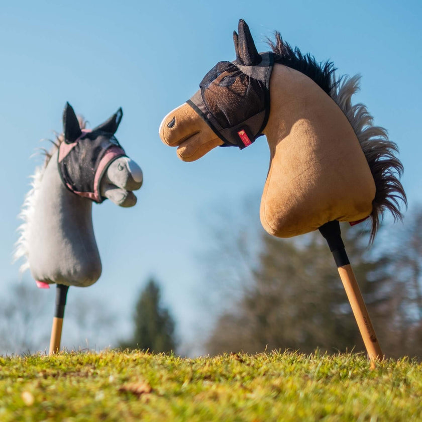 Zwei Hobby Horse Spielzeuge mit Plüschköpfen, eines grau und eines braun, stehen aufrecht auf grasbewachsenem Boden vor klarem blauem Himmel und verschwommenen Bäumen. Beide Pferde tragen kleine Zaumzeuge aus Stoff.