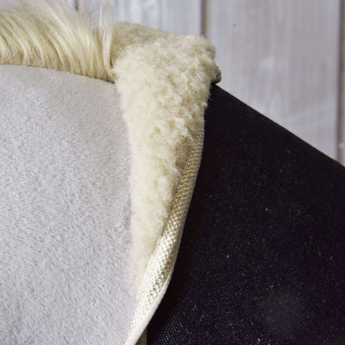 Nahaufnahme der Satteldecke „Set Blue Denim“ von Hobby Horse LarDen mit weicher, flauschiger weißer Schaffellstruktur auf dem Rücken eines dunklen Pferdes, mit Holzwand im Hintergrund.