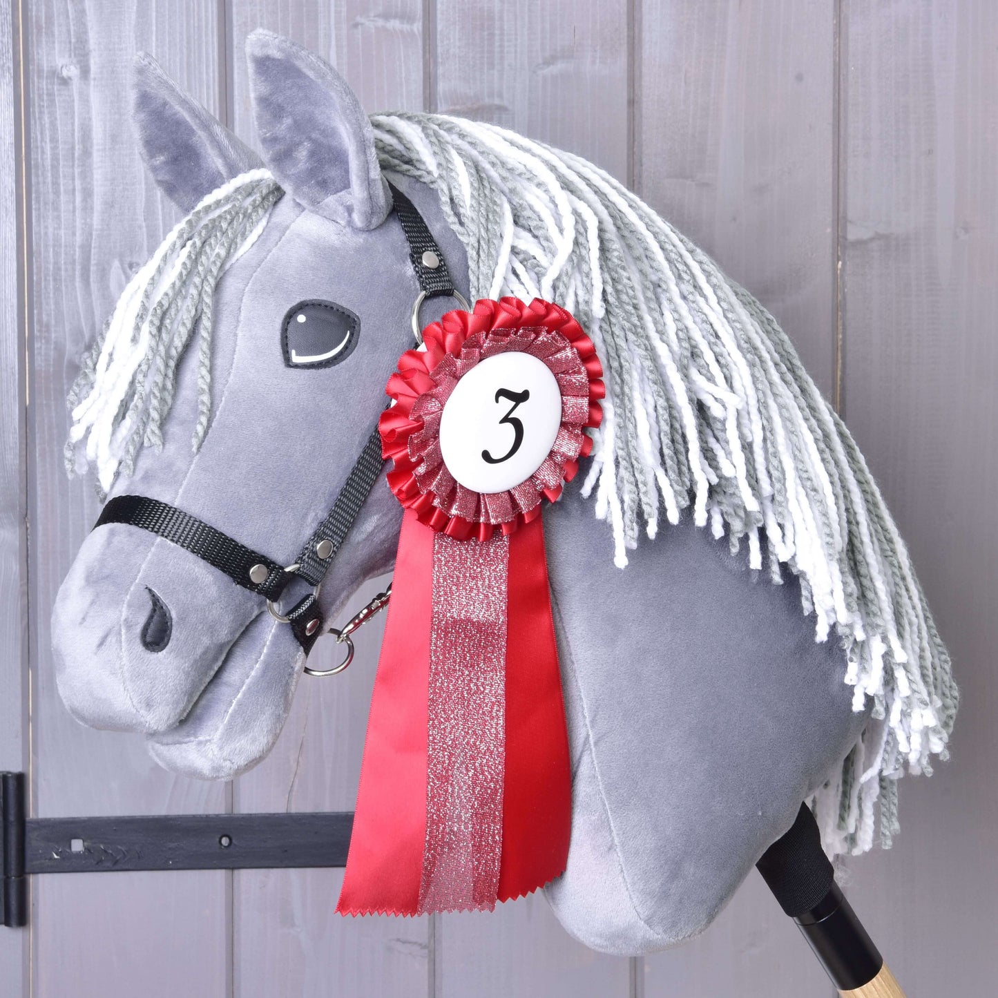 Hobby Horse gris en peluche avec crinière blanche en fil, bride noire et ruban rouge et blanc de l’Ensemble de rubans Hobby Horse NOUVEAU de Hobby Horse LarDen, épinglé avec le numéro 3, devant des panneaux en bois gris.