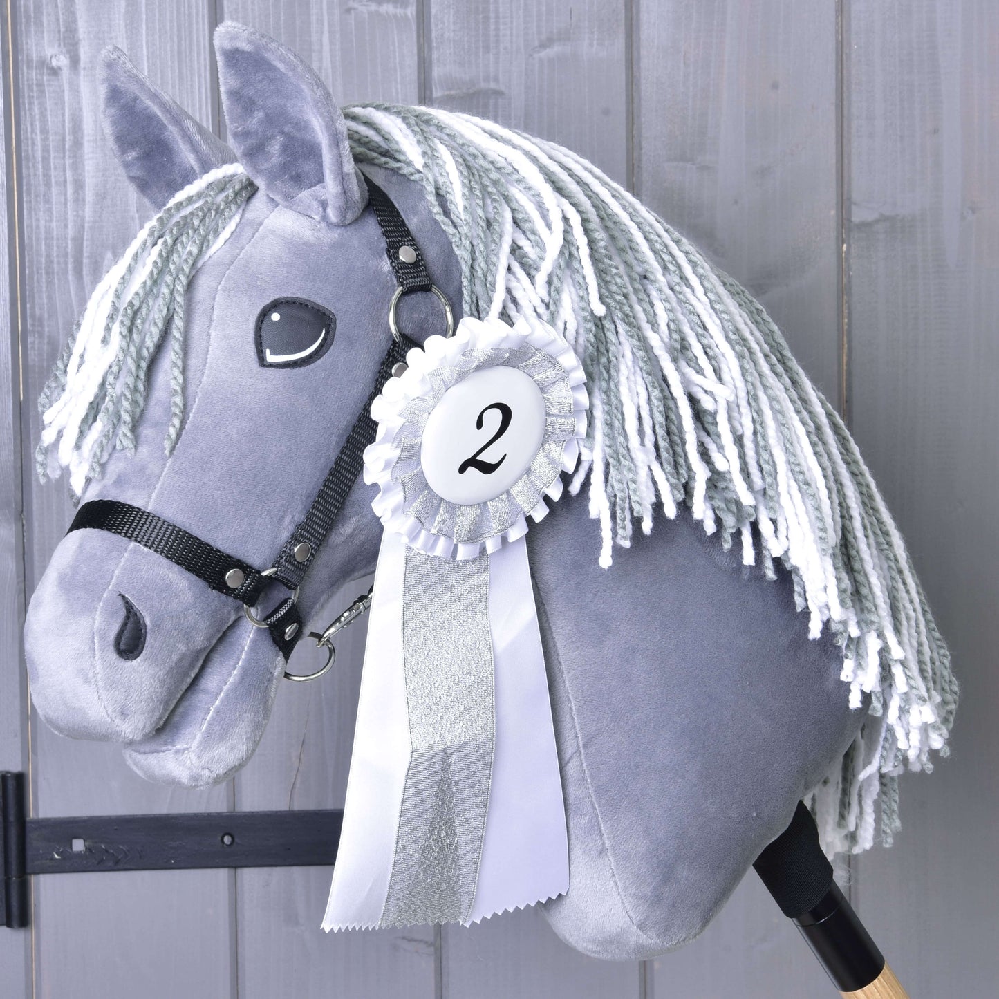 Hobby Horse gris avec crinière blanche en fil et bride noire sur un bâton en bois, présentant un ruban de l’Ensemble de rubans Hobby Horse NOUVEAU (deuxième place, bordure argentée, numéro 2) de Hobby Horse LarDen, devant des planches en bois grises.