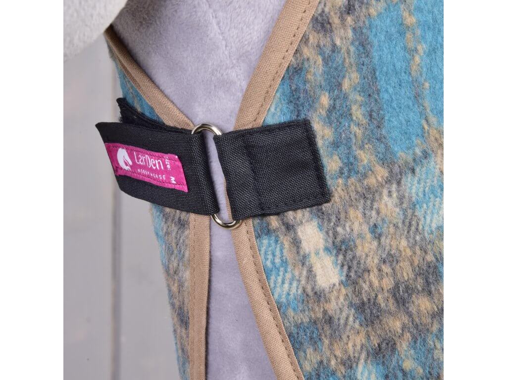 Gros plan sur la couverture d'écurie écossaise à carreaux de Hobby Horse LarDen, avec motifs bleus, marron et gris et bordure beige. Maintenue par une sangle noire avec anneau argenté et finie par une étiquette rose avec logo cheval.