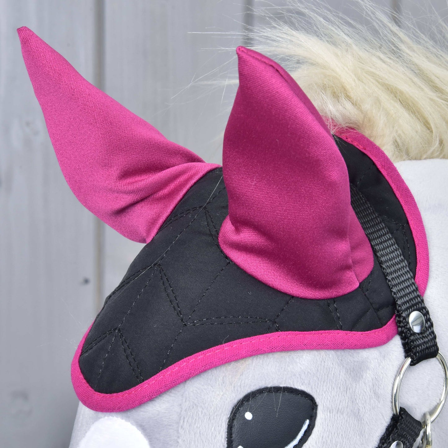 Nahaufnahme einer Hobby Horse LarDen Ohrenkappe Thermo Pinkie in Schwarz und Magenta mit zwei spitzen magentafarbenen Ohren und Steppmuster, die Insektenschutz bietet, vor hellem Holz-Hintergrund.