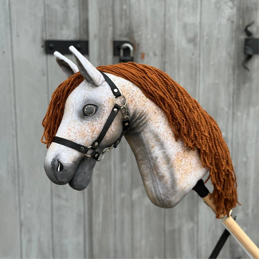 Hobby Horse Pompo mit schwarzem Halfter von Hobby Horse LarDen mit grau gesprenkeltem Gesicht, brauner Garnmähne und schwarzem Zaumzeug, inspiriert vom Hannoveraner Hengst, vor einem verwitterten Holzzaun.