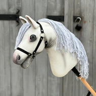 Hobby Horse Atlas mit schwarzem Halfter von Hobby Horse LarDen mit handgefertigtem weißem Stoffkopf, grauer Schnauze, schwarzem Halfter und langer weißer Garnmähne vor einer verwitterten Holztür mit schwarzen Metallscharnieren.