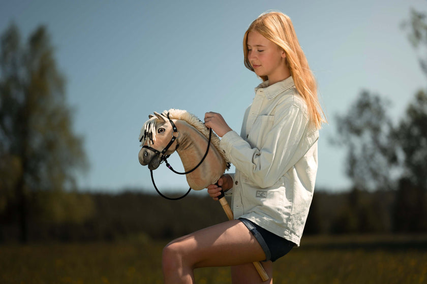 Eine junge Person mit langen blonden Haaren, heller Jacke und Shorts genießt Hobby Horsing mit einem Hobby Horse im Freien auf einem sonnigen, grasbewachsenen Feld mit Bäumen im Hintergrund.