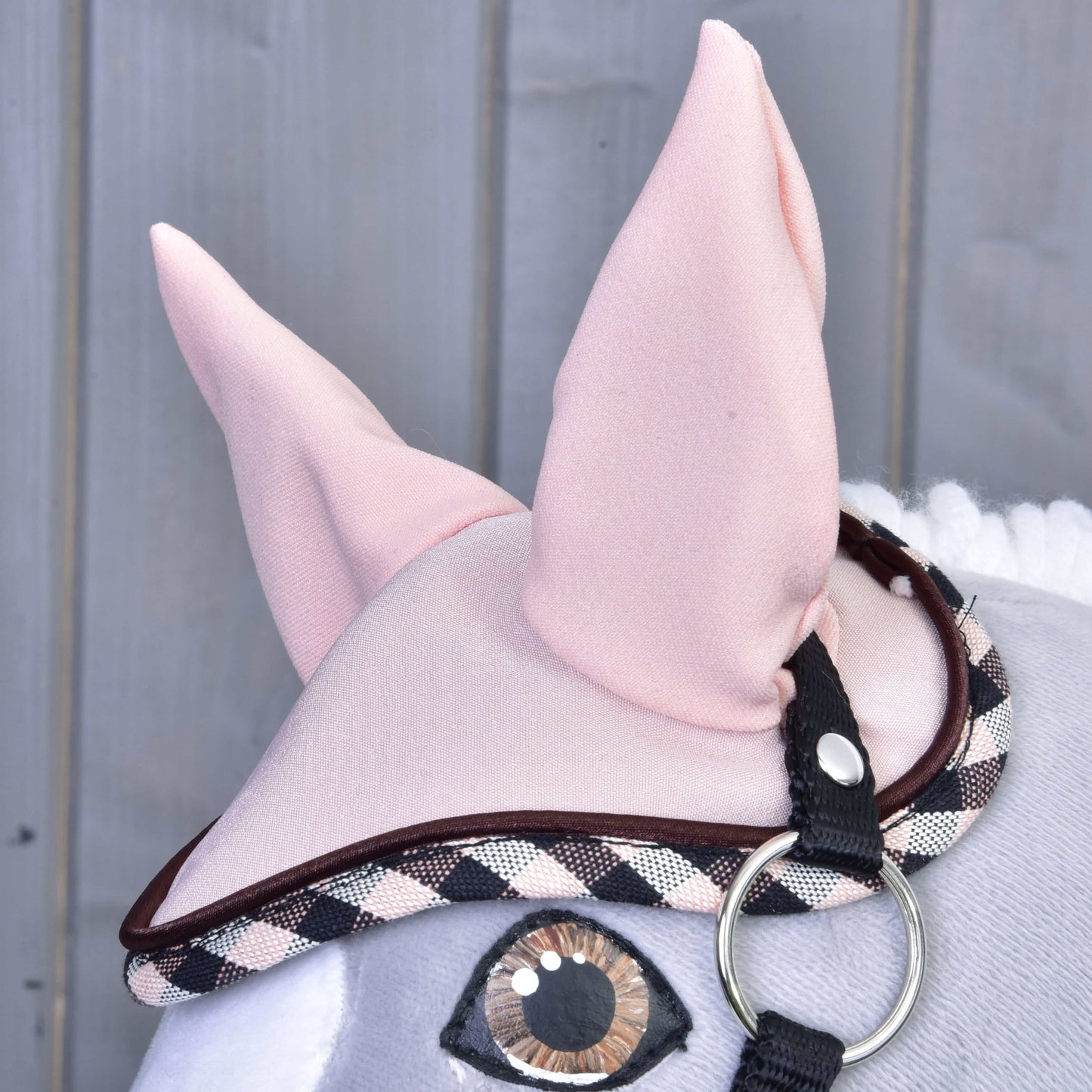 Gros plan sur la peluche Ensemble Abby de Hobby Horse LarDen portant un bonnet d’oreilles rose avec bordure écossaise et sangle noire. Grandes oreilles en tissu et œil cousu visibles sur fond en bois.