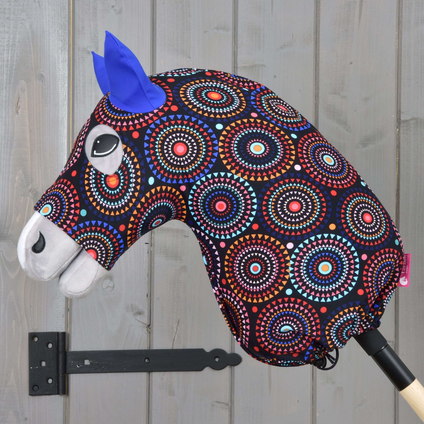 Ein lebhaftes Hobby Horse mit gemustertem Kopf aus Lycra-Stoff, blauen Filzohren und weißen Details auf einem Holzstab – perfekt kombiniert mit dem Hobby Horse LarDen „Hobby Horse Schutzanzug Alice (Lycra)“.