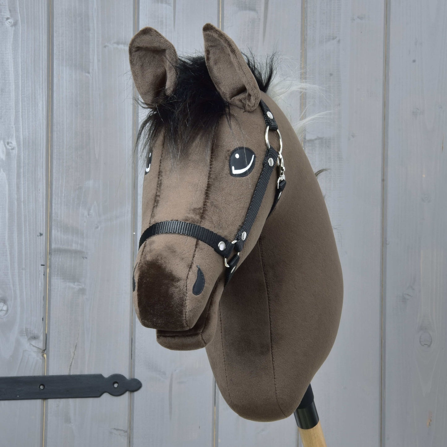 Hobby Horse Alfredo avec licol noir de Hobby Horse LarDen, avec tête d’étalon pur-sang anglais brune en peluche, crinière noire et bride, monté sur un bâton en bois devant un mur en panneaux de bois gris.