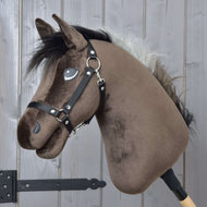 Hobby Horse Alfredo mit Schwarzem Halfter von Hobby Horse LarDen mit braunem Plüschkopf im Stil eines Vollbluts, schwarzer Mähne, weißem Schopf und schwarzem Halfter, auf einem Holzstab vor grauem Holz-Hintergrund.