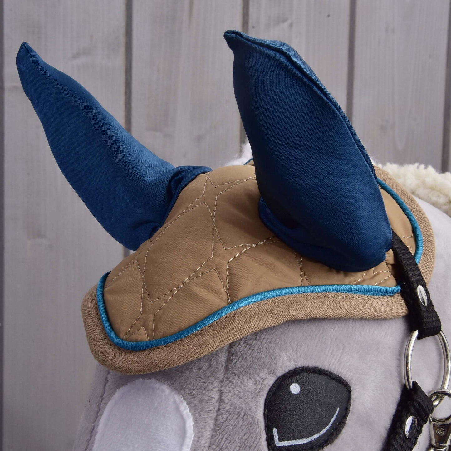 Gros plan de l’Ensemble Étoile Amande de Hobby Horse LarDen montrant son bonnet bleu, sa couverture matelassée beige avec bordure bleue et sa bride sur une tête de Hobby Horse en peluche, sur fond en bois clair.