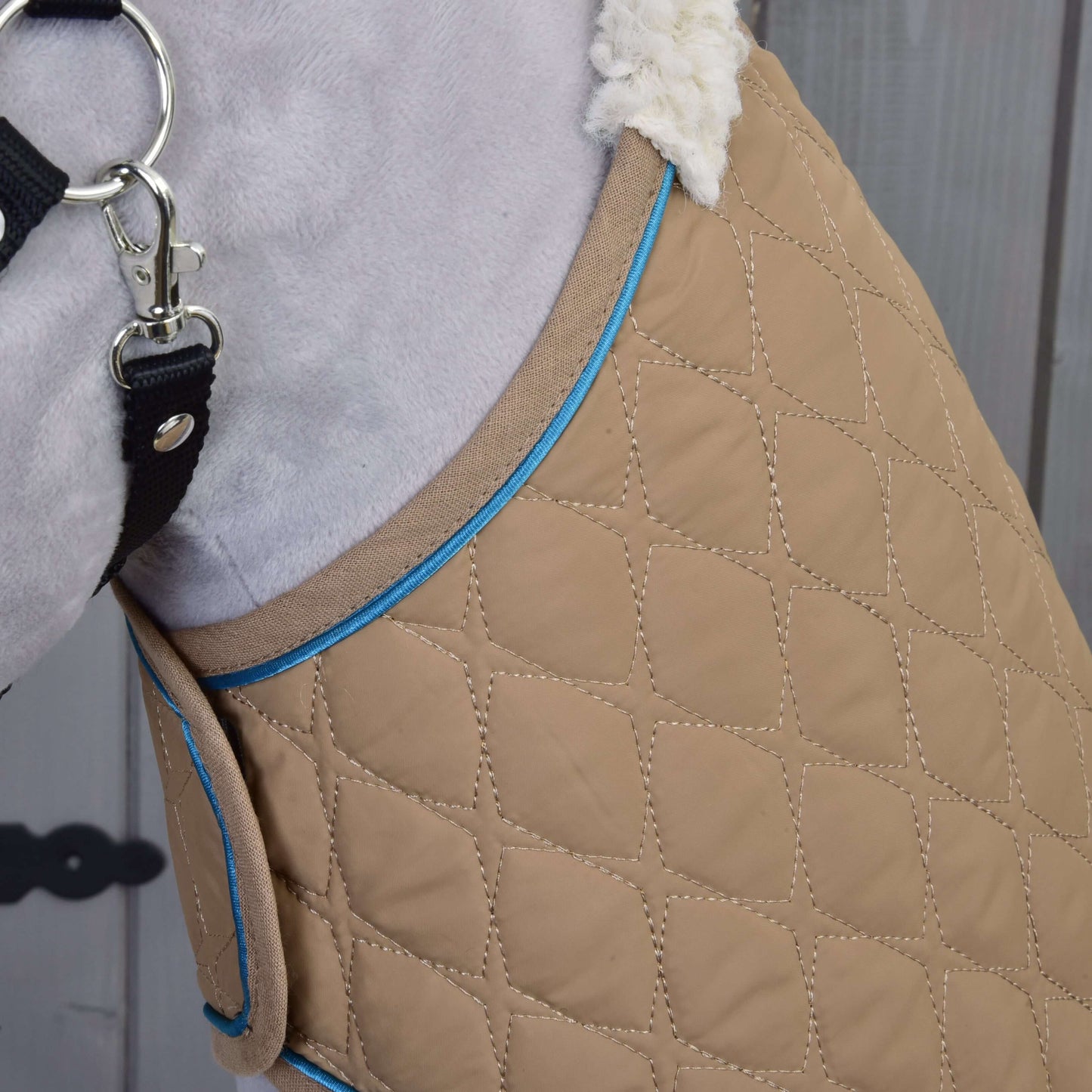 Gros plan du manteau pour chien Ensemble Étoile Amande de Hobby Horse LarDen, avec tissu matelassé beige, bordure bleue, doublure blanche moelleuse, et fermeture par sangle noire et mousqueton métallique, devant un fond en bois.