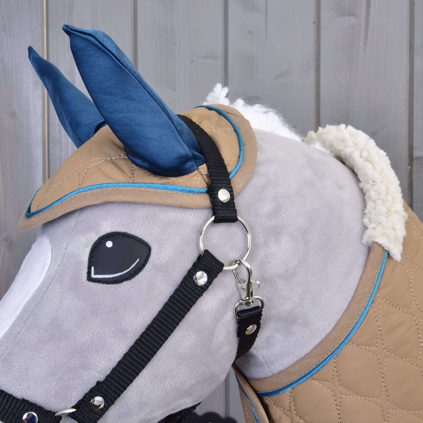 Gros plan de l’Ensemble Étoile Amande de Hobby Horse LarDen, un Hobby Horse en peluche grise avec oreilles bleues, bride noire et couverture matelassée beige et bleue, devant un fond en bois.