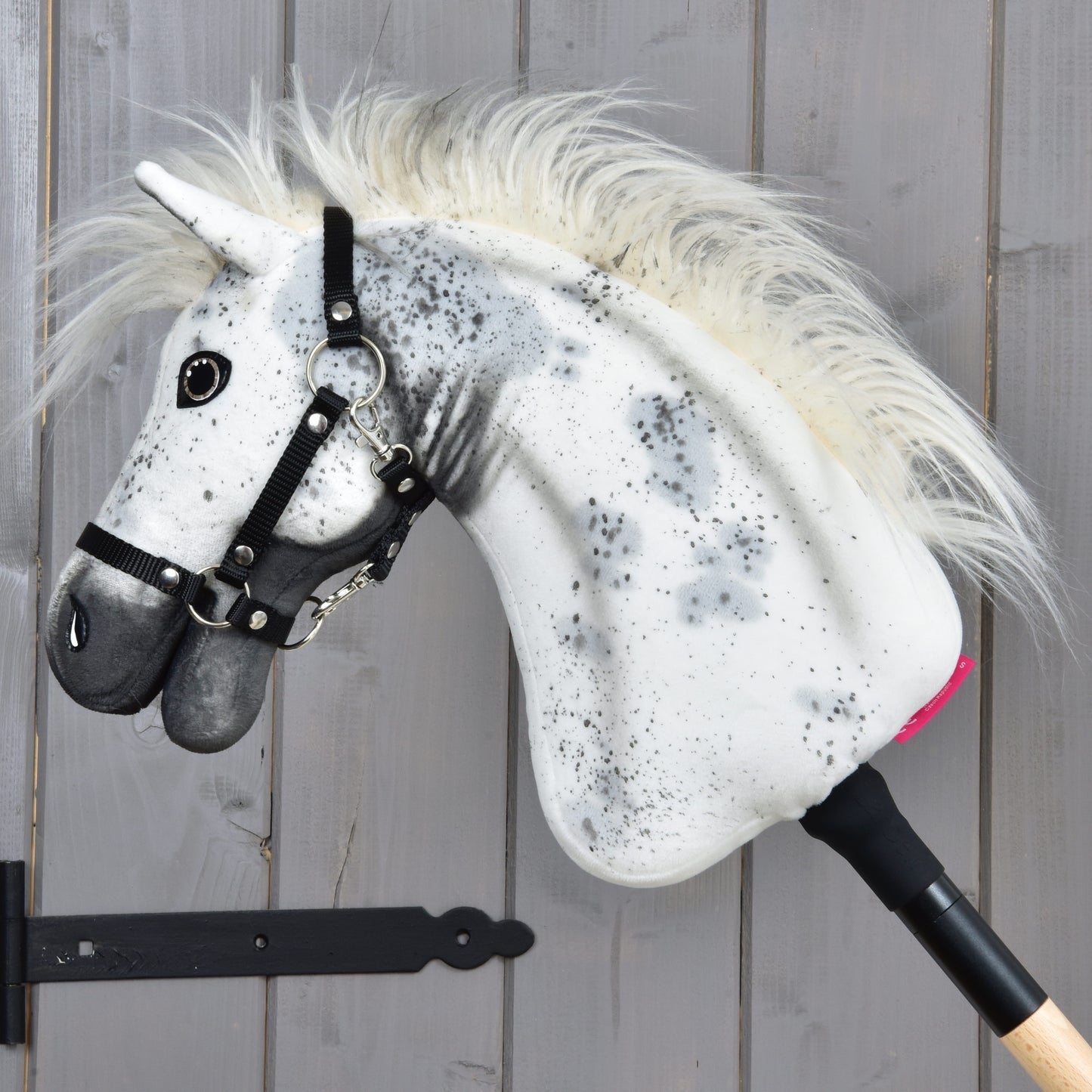 The Hobby Horse LarDen "Hobby Horse Aspen with Black Halter," size S, features a white plush head adorned with black spots, a fluffy mane, and a black halter, all mounted on a wooden stick and displayed against a gray wooden wall.
