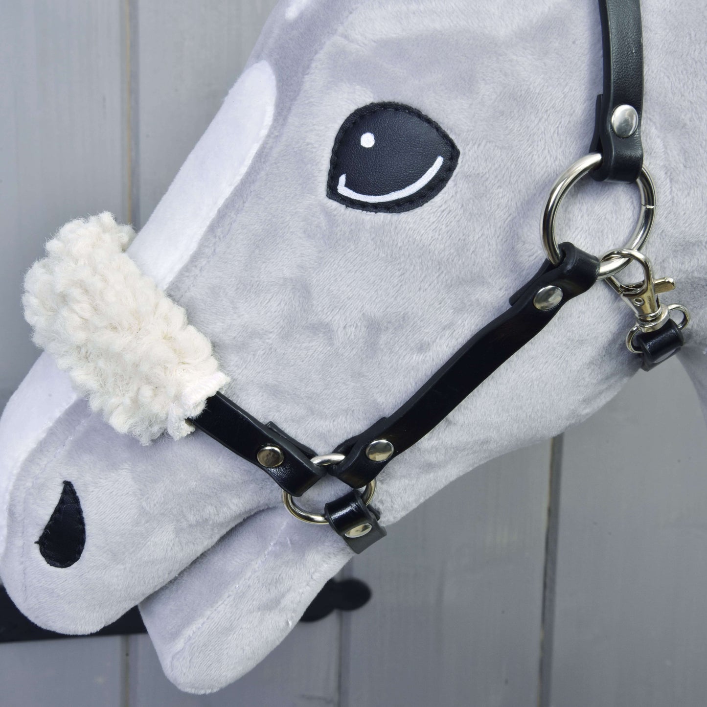 Nahaufnahme des grauen Set Basic Hobby Horse-Kopfes von Hobby Horse LarDen mit schwarzem Zaumzeug mit Zügeln und weißem flauschigem Nasenriemen, vor hellem Holz-Hintergrund.