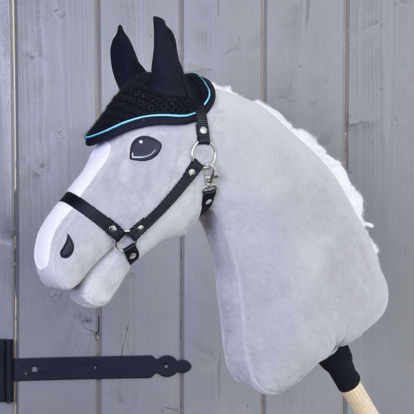 Die Ohrenkappe gehäkelt Blackie von Hobby Horse LarDen ist auf einem grauen Hobby Horse mit schwarzem Zaumzeug zu sehen, auf einem Holzstab montiert und vor einer grauen Holzwand.
