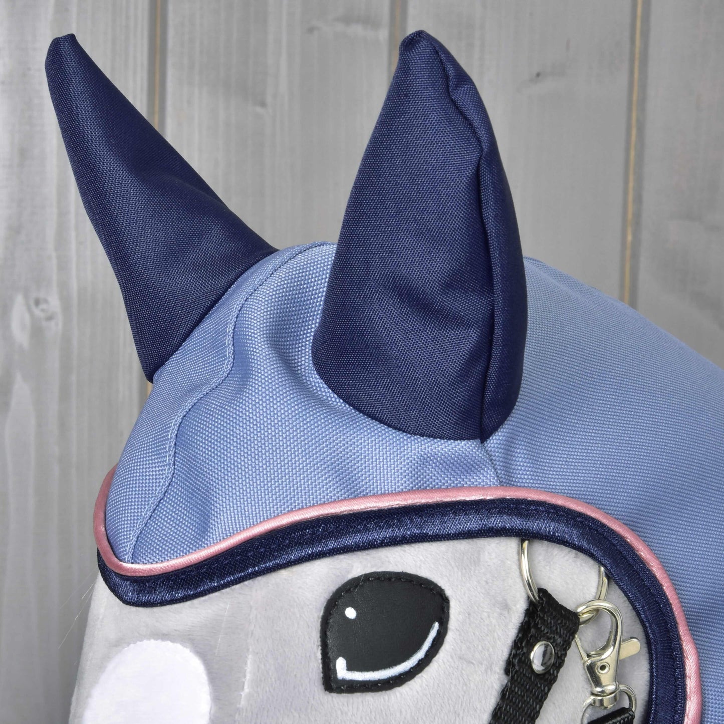 Gros plan de l’Imperméable Bloom de Hobby Horse LarDen, peluche cheval portant un bonnet d’oreilles bleu clair et bleu marine avec grandes oreilles bleu marine et coutures détaillées. Idéal pour présenter des accessoires pour Hobby Horse sur fond en bois.