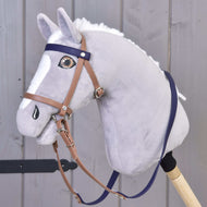 A Hobby Horse LarDen plush hobby horse with light gray fur, white mane and blaze, wearing a Complete bridle with noseband Brown Purple, mounted on a wooden stick against a gray wooden background.