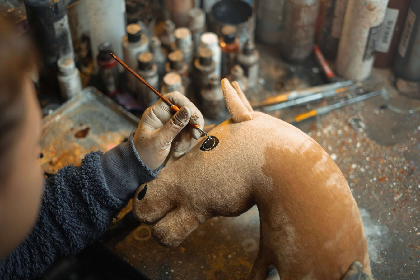 Eine Person mit Handschuhen bemalt das Auge eines braunen Hobby Horse Plüschspielzeugs mit einem feinen Pinsel, umgeben von verschiedenen Farbflaschen und Malutensilien auf einer unordentlichen Werkbank.