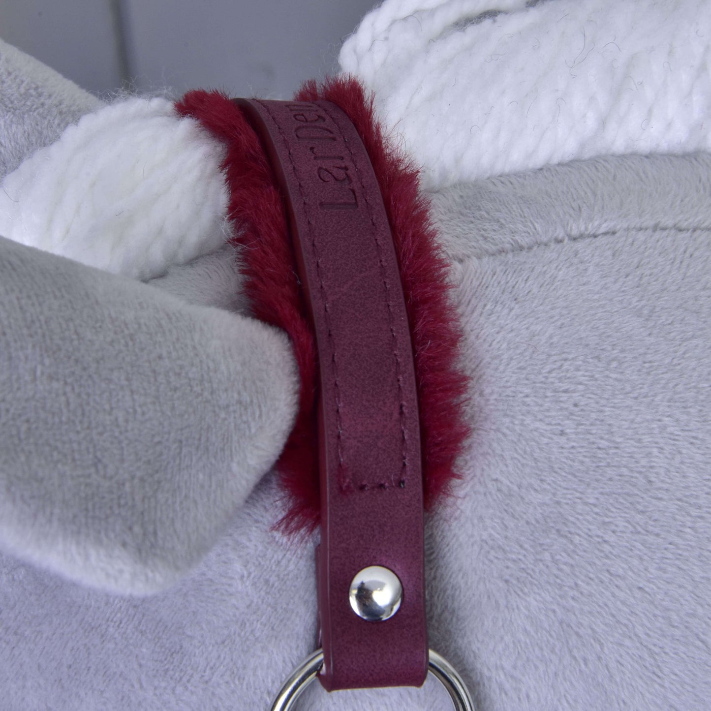 Gros plan sur un tissu en peluche gris avec une corde blanche et une sangle de licol en similicuir bordeaux aux bords rouges duveteux, fixée par des éléments métalliques argentés, un élégant licol avec fourrure - similicuir Bourgogne de Hobby Horse LarDen.