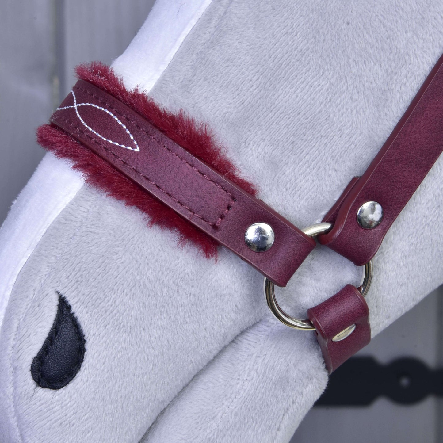 Gros plan sur la tête d’un cheval en peluche gris clair portant le licol avec fourrure - similicuir Bourgogne de Hobby Horse LarDen. Il présente un similicuir rouge rembourré bordeaux et une pièce de naseau noire en forme de goutte. Idéal comme accessoire élégant pour Hobby Horse. Arrière-plan flou.