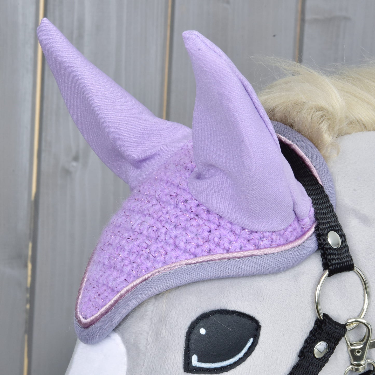 Nahaufnahme eines grauen Plüschpferd-Spielzeugs von Hobby Horse LarDen mit Ohrenkappe Orchid zum Schutz vor Insekten und schwarzem Zaumzeug, vor einem Holzbackground.