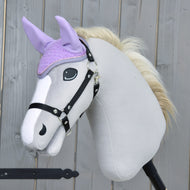 Ein Plüsch-Hobby Horse mit hellgrauem Körper, schwarzem Zaumzeug und cremefarbener Mähne trägt die Hobby Horse LarDen Ohrenkappe Orchid zum Schutz vor Insekten. Im Hintergrund ist ein Holzzaun zu sehen.