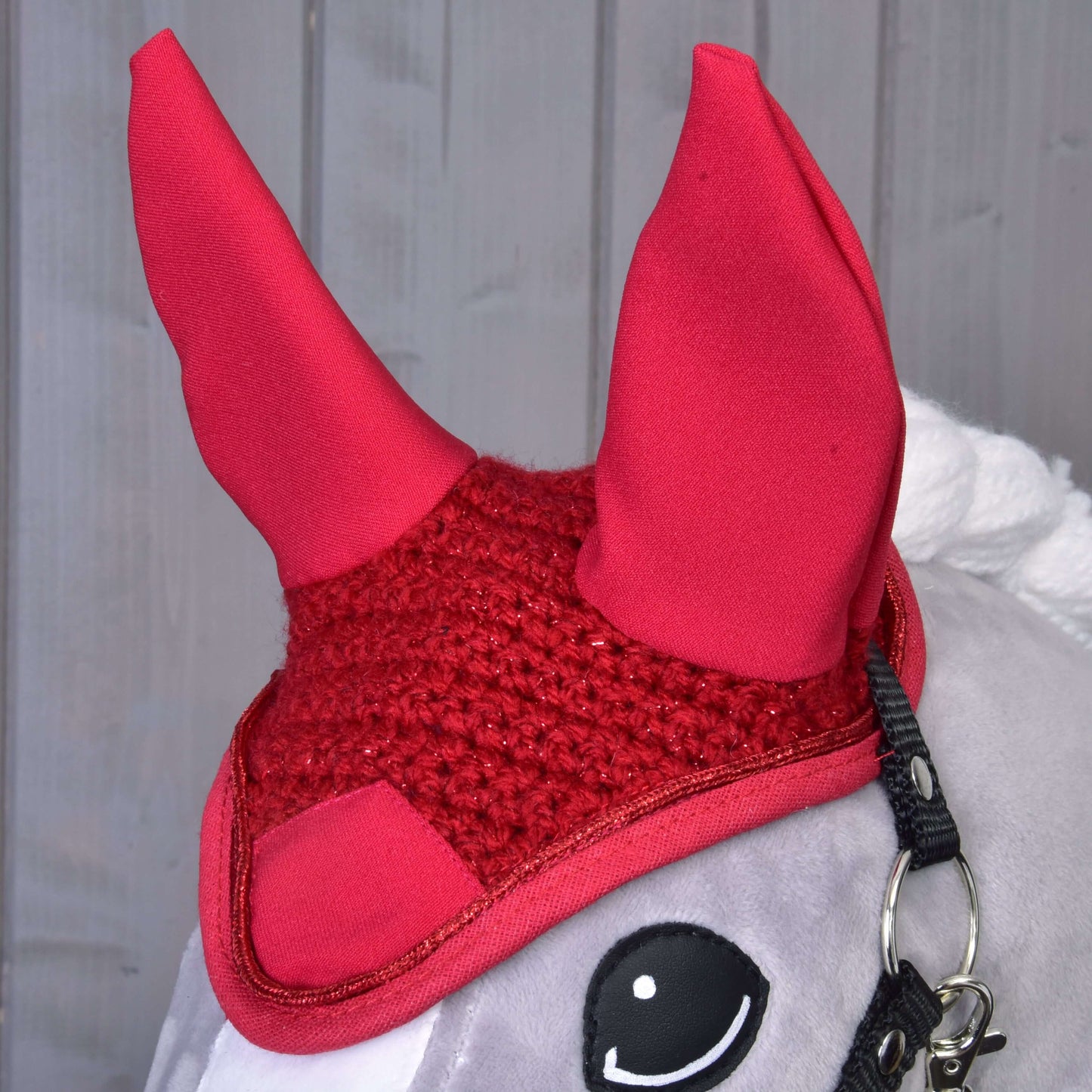 Nahaufnahme eines grauen Plüschpferds mit der Hobby Horse LarDen Ohrenkappe gehäkelt rot mit hohen, spitzen Ohren, die stilvollen Fliegenschutz bietet, vor hellem Holz-Hintergrund.