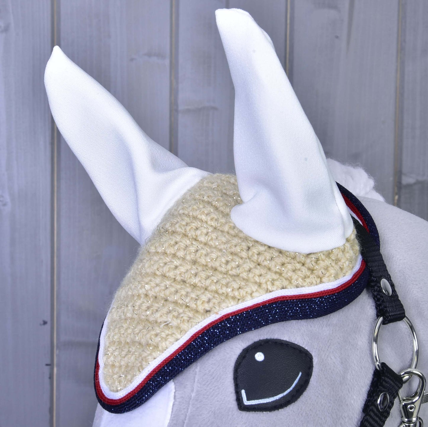 Nahaufnahme eines Pferdekopfes mit der Hobby Horse LarDen Ohrenkappe gehäkelt Nicol in Beige mit weißen Ohren, blau-rotem Besatz und schwarzem Halfter vor grauem Holz-Hintergrund.