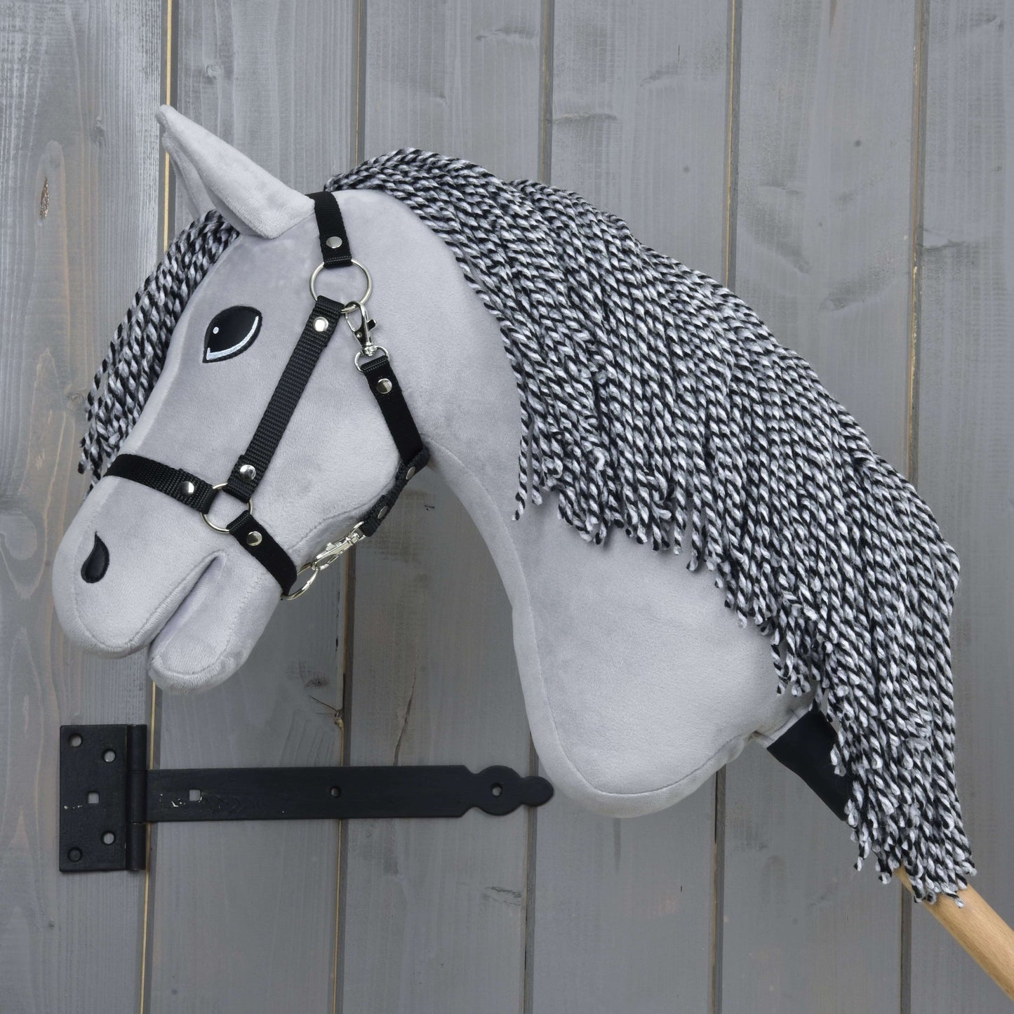 Hobby Horse César avec licol noir de Hobby Horse LarDen, avec tête grise en peluche, crinière en fil noir et blanc, licol noir et grands yeux style dessin animé sur un bâton en bois, parfait pour le jeu imaginatif.