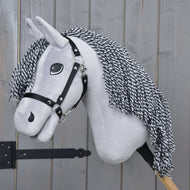 Hobby Horse Caesar mit Schwarzem Halfter von Hobby Horse LarDen mit grauem Plüschkörper, schwarz-weißer Garnmähne und schwarzem Halfter, auf einem Holzstock vor einem Holzzaun-Hintergrund.