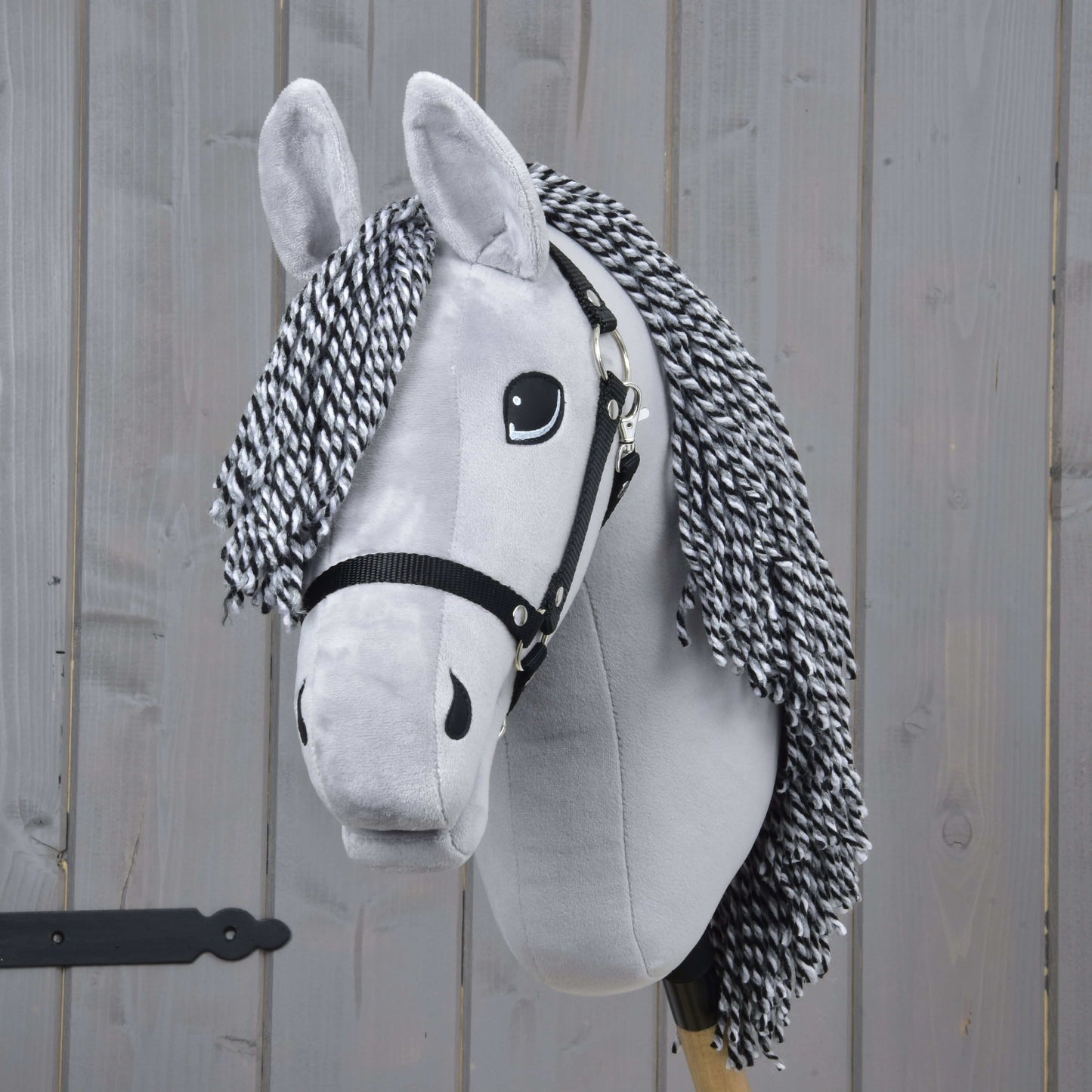 Hobby Horse César avec licol noir de Hobby Horse LarDen, avec corps gris en peluche, crinière en fil noir et blanc, licol noir et yeux expressifs, présenté sur fond de planches en bois.