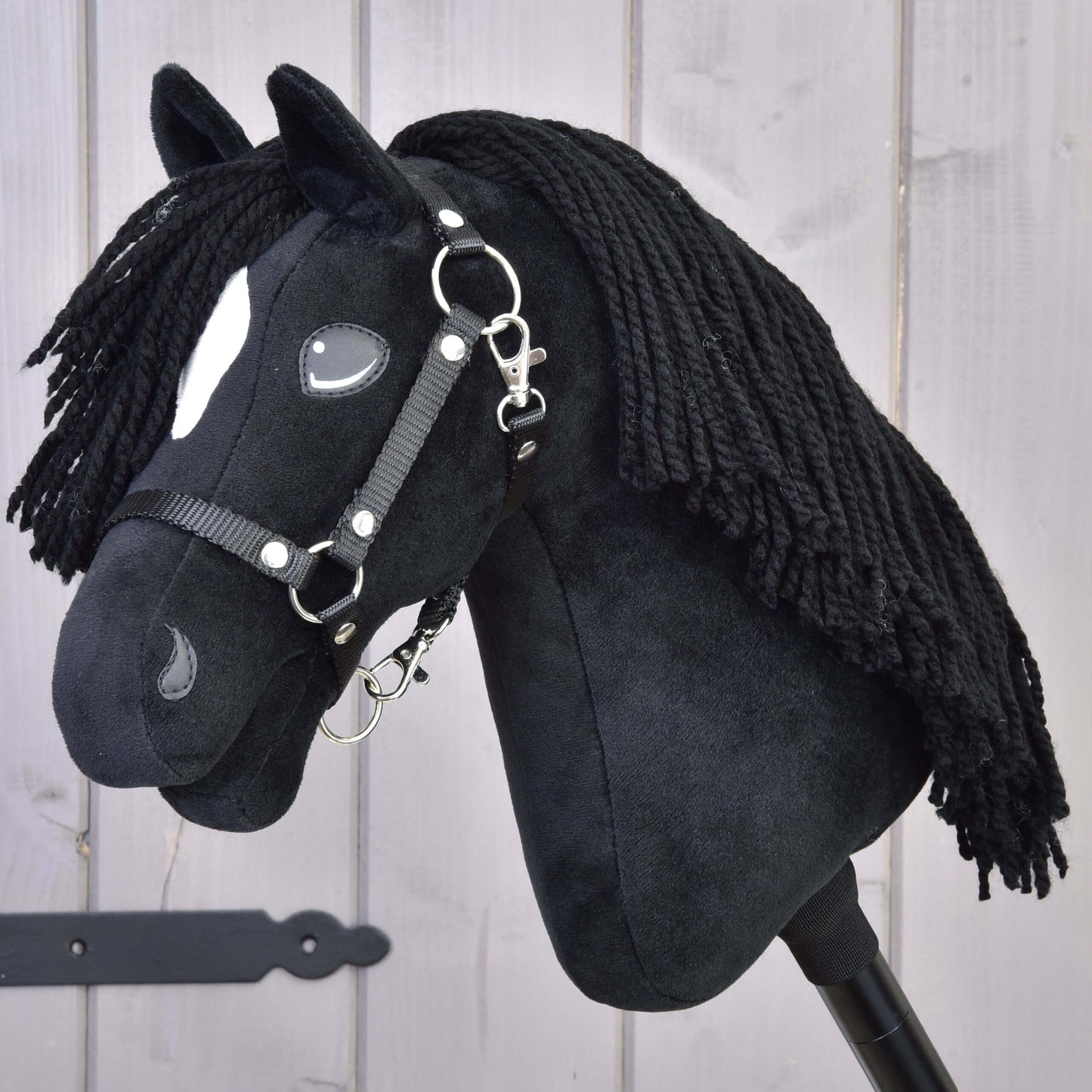 Hobby Horse Cloony avec licol noir de Hobby Horse LarDen, modèle noir en peluche inspiré d'un étalon American Quarter Horse, avec une tache blanche près de l'œil, une crinière noire en fil et un licol noir, présenté sur un fond en bois clair.